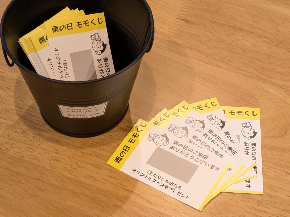 黒い容器に入った黄色い「雨の日 モモくじ」と、テーブルに並べたくじ
