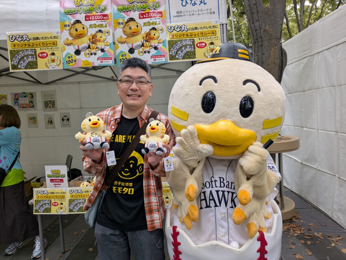 ひな丸のぬいぐるみを両手に持ち、ひな丸と並ぶモモタロ店主